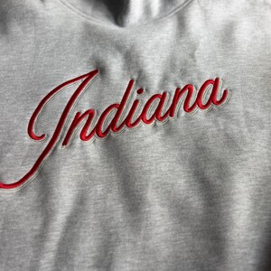 May include: A light grey mock neck sweatshirt featuring the word Indiana embroidered in red cursive script with a white outline. The fabric has a heathered texture, and the embroidery is raised, creating a dimensional effect on the chest area.