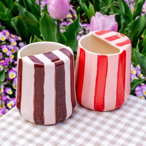 May include: Two ceramic containers with vertical stripes. One has alternating stripes of light pink and brown, the other has stripes of light and dark red. Both have a curved opening at the top. The background features flowers and greenery.