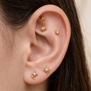 May include: A set of gold-toned star-shaped earrings featuring clear, sparkling stones. The collection includes a dangling star charm, a small star stud, and a multi-star cluster stud, all designed for multiple ear piercings in a bright gold finish.