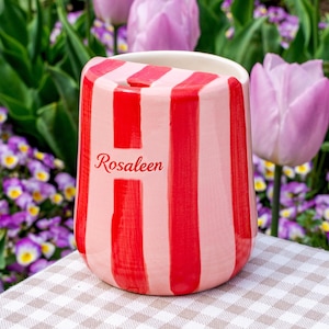 Könnte beinhalten: Ein Keramikbecher mit vertikalen roten und hellrosa Streifen. Der Name Rosaleen ist in roter Schreibschrift auf der Vorderseite aufgedruckt. Das Gefäß hat eine glatte, glänzende Oberfläche und eine leicht geschwungene Form.