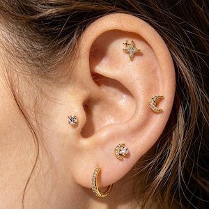 May include: A set of five gold-tone earrings featuring celestial motifs. The collection includes a four-pointed star stud with clear stones, a crescent moon stud, a small round solitaire stud, a moon and star stud, and a small hinged hoop earring.