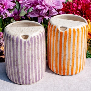 Peut inclure: Deux vases muraux en céramique. L'un est blanc avec des rayures verticales violettes, l'autre est blanc avec des rayures verticales orange. Les deux ont une finition mouchetée et un dessus arrondi avec un trou.