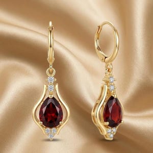 Può includere: Un paio di orecchini pendenti color oro. Ogni orecchino presenta una gemma rosso scuro a forma di goccia, impreziosita da piccoli cristalli trasparenti. Gli orecchini hanno una chiusura a leva.