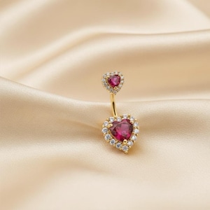 May include: A gold-coloured belly button ring featuring heart-shaped ruby-red gemstones. Each gemstone is encircled by a halo of small, clear crystals. The ring is set against a satin background.