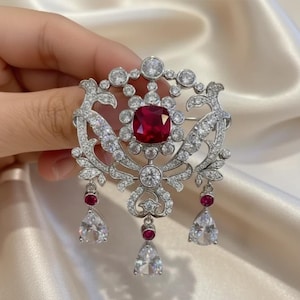 May include: A silver-toned brooch with a large, square, red gemstone at the center, surrounded by smaller clear stones. The brooch has an ornate, floral design with dangling teardrop-shaped clear stones and smaller red gems.