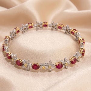 May include: A floral-inspired bracelet featuring alternating ruby gemstones and flower-shaped accents. The bracelet has a silver and gold-tone metal setting, with small yellow and clear accent stones. The bracelet is displayed on a cream-colored satin surface.
