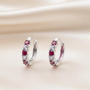 May include: Silver hoop earrings, each featuring alternating red and clear gemstones. The earrings are displayed against a soft, neutral backdrop. The gemstones are evenly spaced around the hoops, creating a striking contrast.