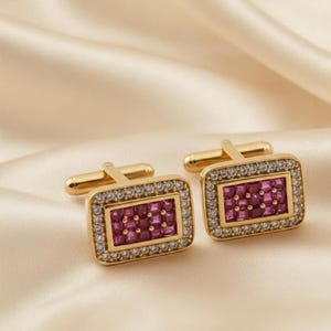 May include: A pair of gold cufflinks with a rectangular design. Each cufflink features a central section of small, square, ruby-coloured stones, surrounded by a border of clear, sparkling crystals. The cufflinks are set against a soft, cream-coloured background.