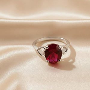 Peut inclure: Une bague en argent avec une grosse pierre rouge foncé de forme ovale. La bague présente des accents en forme de cœur de chaque côté de la pierre. La bague est posée sur un fond crème clair.