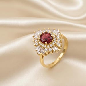 Puede incluir: Anillo dorado con una piedra roja oscura central, rodeada de pequeñas piedras claras con corte baguette. El diseño del anillo presenta un patrón de rayos de sol alrededor de la piedra central, sobre un fondo crema claro.