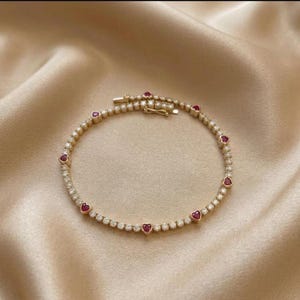 May include: A gold-toned bracelet with a delicate design. The bracelet features small, round, clear stones and heart-shaped red stones. The bracelet is set against a satin background.