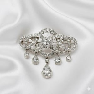 Puede incluir: Un broche plateado con un diseño intrincado de bucles entrelazados y gemas claras y brillantes. El broche presenta una gran piedra central y varios colgantes en forma de pera, sobre un fondo de satén blanco.