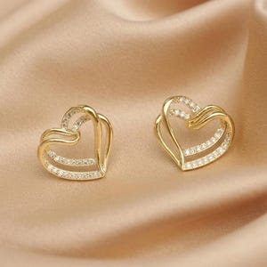 May include: A pair of gold-toned heart-shaped stud earrings. Each earring features an open-work design with two overlapping heart outlines, where the lower heart section is encrusted with small, clear, sparkling stones along the front surface.