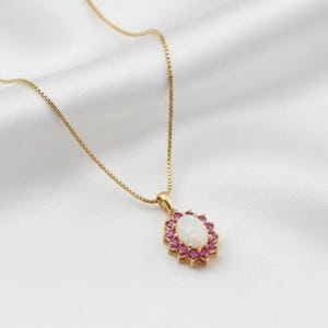 Puede incluir: Collar dorado con colgante. El colgante presenta una ópalo blanco ovalado rodeado de pequeñas piedras rosas. La cadena es delicada, sobre un fondo blanco sedoso.