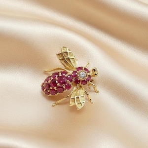 May include: A gold-colored bee brooch with ruby-colored gems forming the body and a central diamond-like gem. The wings and legs are detailed with intricate patterns. The brooch is set against a satin background.