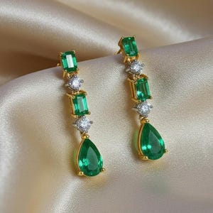 May include: A pair of gold-toned drop earrings featuring emerald-green gemstones and small, round, clear stones. The earrings have a linear design with a pear-shaped emerald at the bottom.