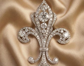 May include: A diamond fleur-de-lis brooch with a detailed design. The brooch features multiple round-cut diamonds set in a silver-colored metal. The design includes a central vertical element with a series of diamonds and curved side elements.