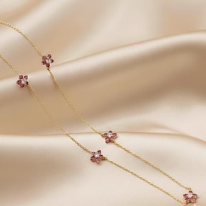 Può includere: Una delicata collana d'oro con piccoli pendenti a forma di fiore. Ogni fiore ha una gemma bianca centrale circondata da petali rosa. La collana è esposta su uno sfondo di raso.