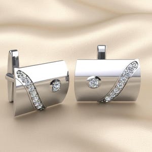May include: A pair of silver cufflinks with a curved row of small, clear gemstones and a central, larger gemstone. The cufflinks have a rectangular shape with rounded edges and a polished finish. The background is a light beige color.