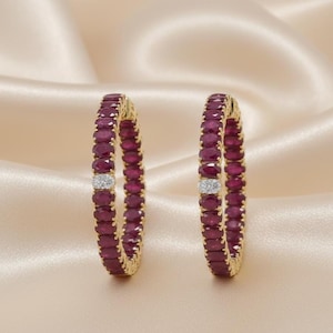 May include: A pair of circular hoop earrings crafted from yellow gold, featuring a continuous row of oval-cut deep red rubies. Each earring is accented with a single small segment of pavé-set white diamonds positioned along the hoop's edge.