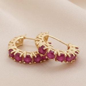 May include: A pair of gold hoop earrings featuring a row of round, deep red rubies set in prong settings. The outer edge of the gold hoops displays a decorative cut-out pattern, and each earring is secured with a thin, straight post backing.