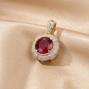 May include: A gold pendant featuring a large, round, deep red gemstone at the center. The stone is surrounded by a thick, textured gold border encrusted with numerous small, clear pavé-set stones. A gold bail with pavé stones sits on top.