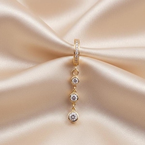 Op de afbeelding: Een goudkleurige oorbel met drie heldere, ronde edelstenen die aan een ring bungelen. De ring is bezet met kleine, heldere edelstenen. De oorbel wordt gepresenteerd op een satijnen stof.