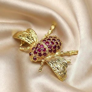 May include: A gold-colored bee brooch with ruby-colored gemstones. The brooch features detailed wings and body segments, set against a satin background. The ruby gemstones are clustered on the bee's body, adding a vibrant contrast to the gold.