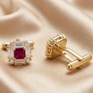 May include: A pair of gold cufflinks with a square design. Each cufflink features a central, square, deep red gemstone surrounded by rectangular and round clear stones. The cufflinks are set against a gold-colored, satin-like background.