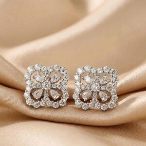 May include: A pair of square-shaped, silver-toned earrings. Each earring features a floral design with clear, sparkling stones. The earrings are set against a soft, golden-colored background.