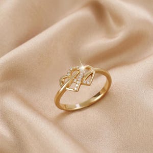 May include: A gold-colored ring with a double heart design. One heart is encrusted with small, clear gemstones. The ring is set against a soft, beige background, creating a delicate and romantic aesthetic.