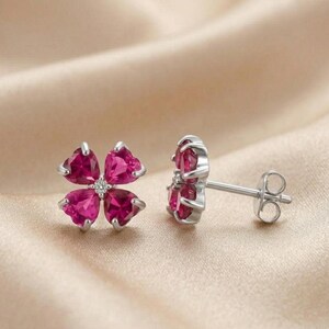 May include: A pair of silver stud earrings. Each earring features a four-petal flower design with heart-shaped, magenta-colored gemstones. A small, clear gemstone is set in the center of each flower.