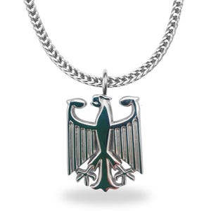 Könnte beinhalten: Silberfarbene Halskette mit einem detaillierten Adler-Emblem. Der Anhänger hängt an einer geflochtenen Kette. Das Adler-Design hat ein strukturiertes Aussehen, mit weit ausgebreiteten Flügeln. Die Halskette wird vor weißem Hintergrund präsentiert.