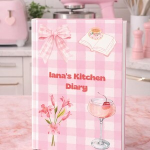 May include: A pink and white checked kitchen diary with the text "Iana's Kitchen Diary". The diary features illustrations of a bow, open book, flowers, and a cocktail. A pink stand mixer and other kitchen appliances are in the background.