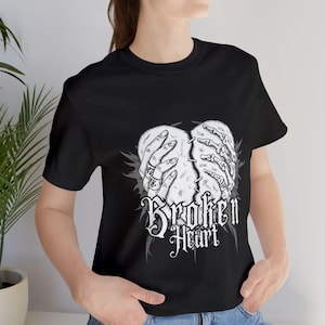 Op de afbeelding: Zwart t-shirt met een zwart-wit afbeelding van een gebroken hart dat door handen wordt vastgehouden. De tekst "Broken Heart" staat in gotische letters.