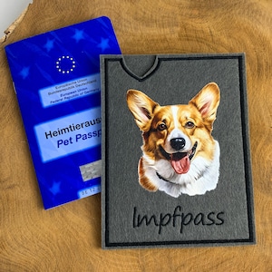 Könnte beinhalten: Eine graue Filzhülle für einen Heimtierausweis mit einem gestickten Porträt eines braun-weißen Corgi-Hundes und dem schwarzen Schriftzug Impfpass. Daneben liegt ein blauer EU-Heimtierausweis mit Sternen und dem Text Pet Passport.