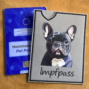 Könnte beinhalten: Ein grauer Filz-Haustierpasshalter mit einer schwarzen Französischen Bulldoggen-Illustration und dem Wort "Impfpass" in schwarzer Stickerei. Daneben ist ein blauer EU-Heimtierausweis zu sehen.