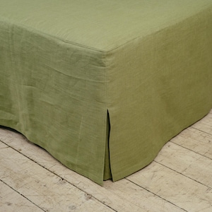 Puede incluir: Primer plano de una falda de cama verde salvia hecha de lino. La falda de la cama tiene un diseño a medida con un detalle de esquina dividida. La tela tiene una apariencia texturizada y un color suave y apagado.