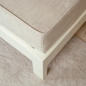 May include: Close-up of a beige upholstered cushion on a white wooden frame. The cushion has a textured appearance and is bordered by a stitched seam. The frame is square and painted white. The corner of the frame and cushion are visible.