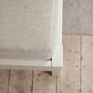 May include: Close-up of a section of a bed frame with a light beige linen upholstered headboard. The frame is white, and the headboard has a textured appearance. The bed frame sits on a light-colored wooden floor.