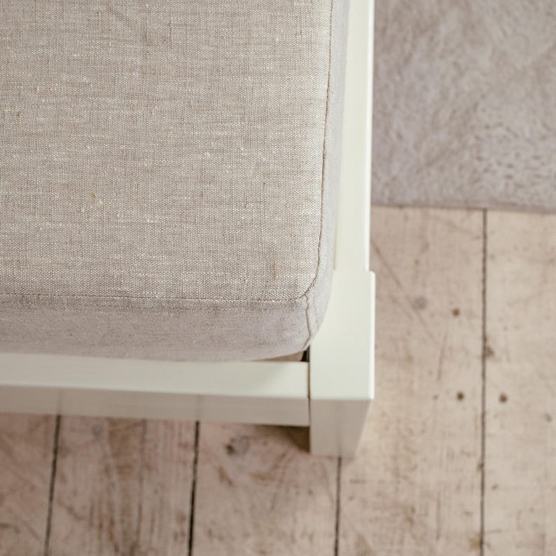 May include: Close-up of a section of a bed frame with a light beige linen upholstered headboard. The frame is white, and the headboard has a textured appearance. The bed frame sits on a light-colored wooden floor.