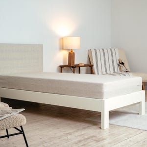 May include: A minimalist bed frame with a neutral-toned mattress and headboard. The bed is set against a white wall, with a small side table and lamp nearby. The bed frame is white, and the mattress is a light beige color.