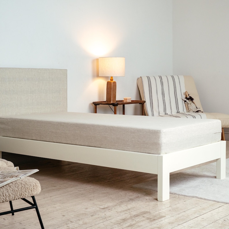 May include: A minimalist bed frame with a neutral-toned mattress and headboard. The bed is set against a white wall, with a small side table and lamp nearby. The bed frame is white, and the mattress is a light beige color.