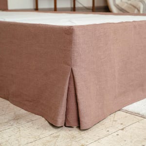 Op de afbeelding: Een stofroze bedrok met een geplooid hoekdetail. De stof heeft een linnen-achtige textuur en valt tot op de vloer. De bedrok is effen van kleur en voegt een vleugje elegantie toe aan het bed.