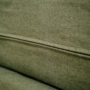 May include: Close-up of olive green canvas fabric with a visible woven texture. The fabric is divided by a seam, creating a subtle geometric pattern. The color is a muted, earthy tone, suggesting a natural material.