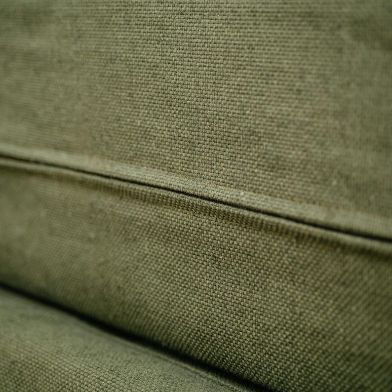 May include: Close-up of olive green canvas fabric with a visible woven texture. The fabric is divided by a seam, creating a subtle geometric pattern. The color is a muted, earthy tone, suggesting a natural material.