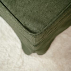 May include: Close-up of a section of a green fabric sofa. The fabric has a textured appearance and is a solid, muted green color. The corner of the sofa is visible, showing the seams and the shape of the cushion.