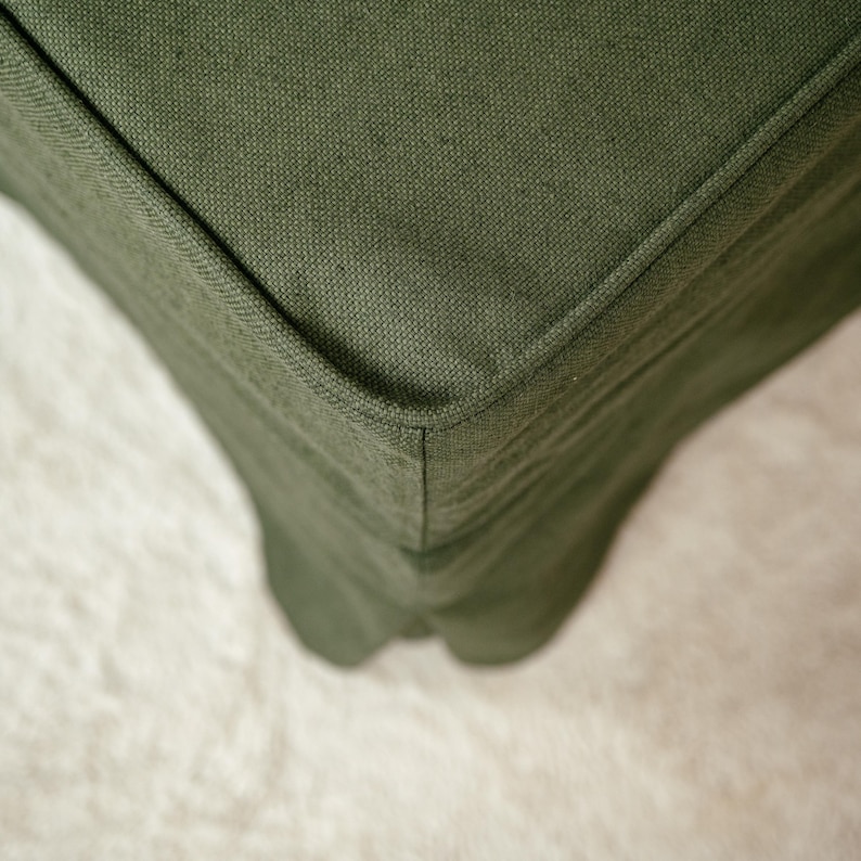 May include: Close-up of a section of a green fabric sofa. The fabric has a textured appearance and is a solid, muted green color. The corner of the sofa is visible, showing the seams and the shape of the cushion.