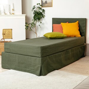 May include: A green upholstered daybed with a matching headboard and skirt. The bed is adorned with three colorful pillows: orange, yellow, and green. The room has a neutral color palette with a white fireplace and a light-colored rug.