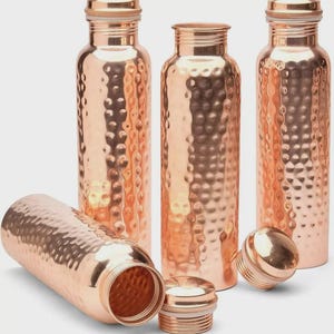 Puede incluir: Botellas de agua de cobre con textura martillada. Las botellas tienen forma cilíndrica con tapón de rosca. Una botella está tumbada de lado, con el tapón quitado. Las botellas son de color cobre metálico brillante.